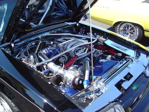 1986 Ford Bronco - The engine bay of a black car features extensive chrome and blue aftermarket parts, with another vehicle in the background.