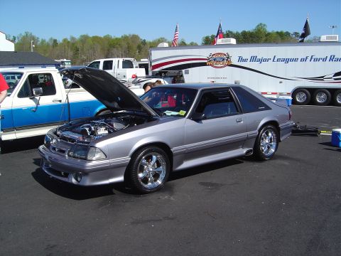 1986 Ford Bronco - A silver coupe, likely a Ford Mustang, is displayed with its hood open, parked near a trailer at an outdoor event.