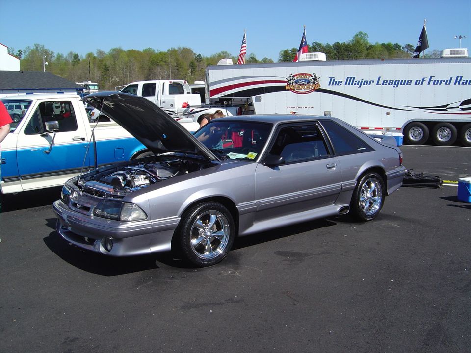 Silver coupe with hood open, chrome wheels, and a trailer in the background