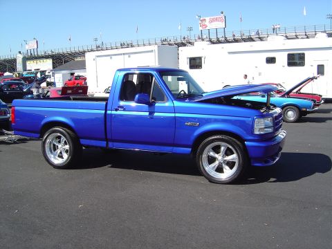 1986 Ford Bronco - A bright blue pickup truck is parked at an outdoor event with its hood open, showing other vehicles and grandstands.