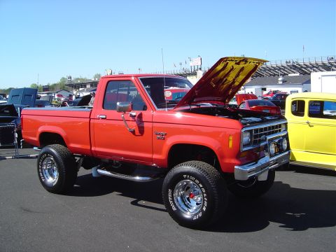 1986 Ford Bronco - A red pickup truck with its hood up is displayed at an outdoor event with other vehicles.