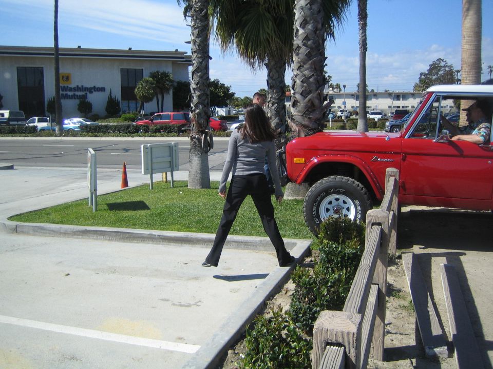 Red Ford Bronco with white top, driver visible. Woman stands in front of it