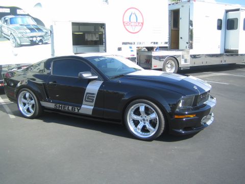 1968 Ford Mustang - A black Shelby Mustang with silver racing stripes is parked in front of a trailer with a Cobra logo.