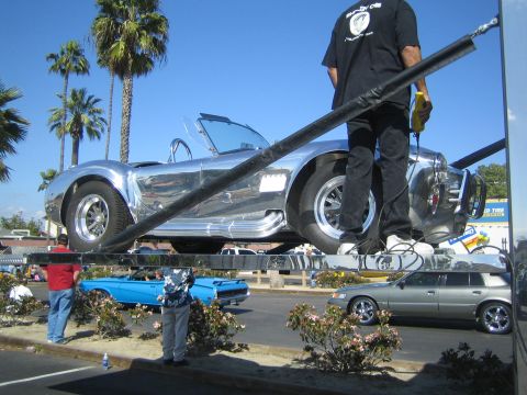 1968 Ford Mustang - A chrome roadster is elevated on a lift, with a man standing nearby. Other vehicles are parked below.