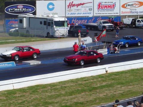 1986 Ford Bronco - Drag racing event with two cars competing on the track, RV and support vehicles visible.