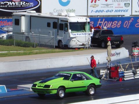 1986 Ford Bronco - A modified 1985 Ford Mustang hatchback is on the drag strip, with an RV and pickup truck in the background.