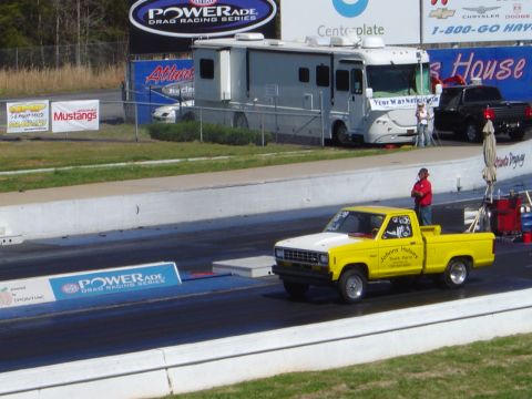 1986 Ford Bronco - A yellow and white pickup truck is on a drag strip, with an RV and another truck in the background.