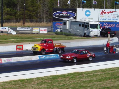 1986 Ford Bronco - Two vehicles compete on a drag strip with an RV and spectator stands in the background.