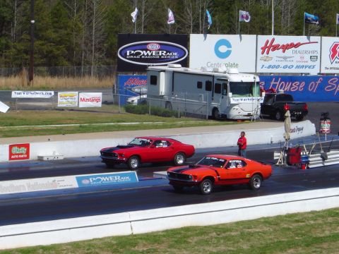 1986 Ford Bronco - Two classic Ford Mustangs compete on a drag strip, with a large RV and support vehicles in the background.