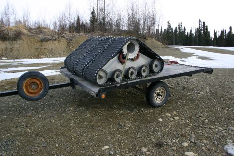 2003 Jeep TJ - A tracked vehicle is transported on a trailer across a gravelly area with patches of snow.