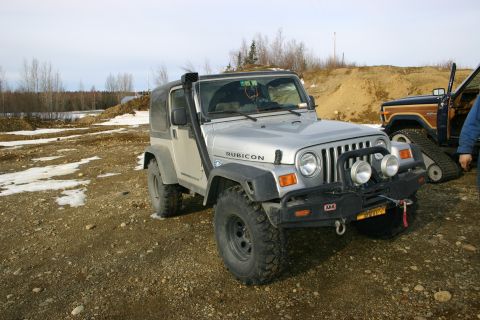 2003 Jeep TJ - A silver Jeep Wrangler is parked on a gravelly surface next to another vehicle with tracks.