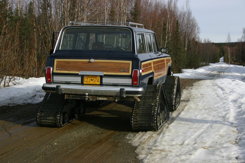 Rear view of a dark blue Jeep Grand Wagoneer with wood paneling, equipped with tracks instead of wheels, on a snowy dirt road