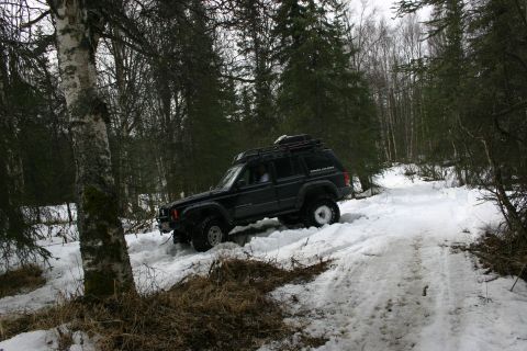 2003 Jeep TJ - A black Jeep Cherokee XJ navigates a snow-covered forest trail, equipped with a roof rack and off-road tires.