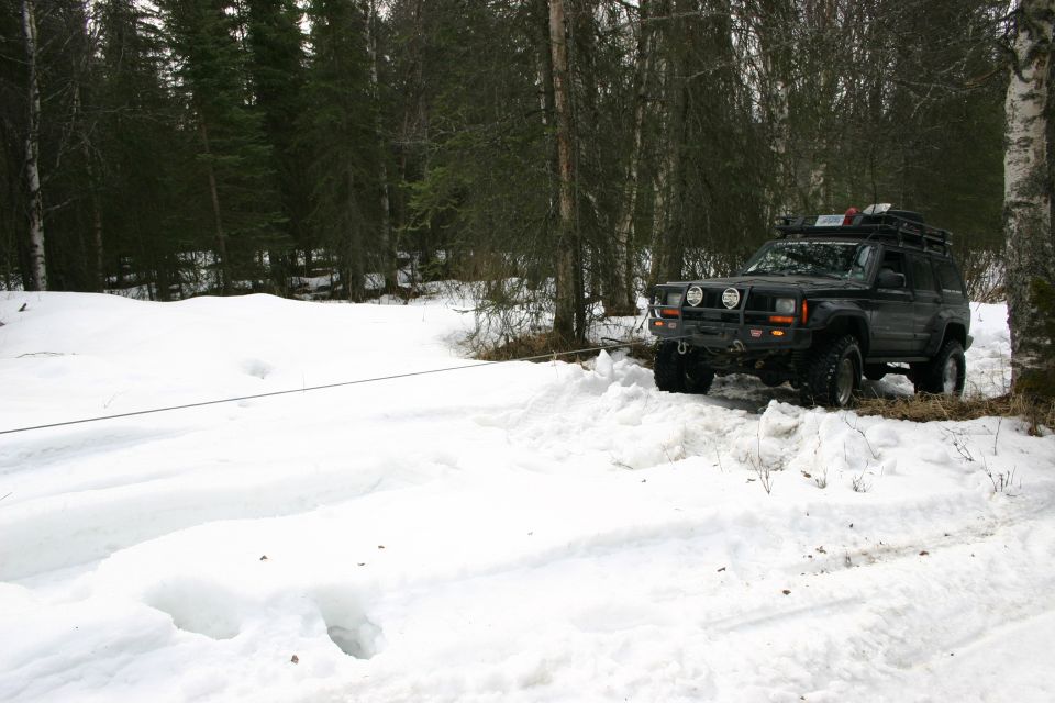 Black Jeep Cherokee XJ with roof rack and winch, angled three-quarters from the front, in a snowy wooded area