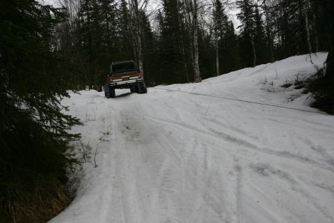 2003 Jeep TJ - A brown SUV with large tires is being towed up a snowy incline by a cable, surrounded by trees.