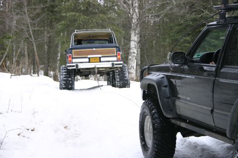 2003 Jeep TJ - A blue Jeep Grand Wagoneer with wood paneling, equipped with snow tracks, is being towed by a black Jeep Cherokee XJ through a snowy forest.
