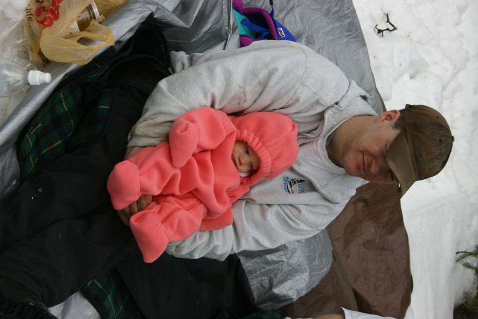 Man in a baseball cap holding a baby in a pink snowsuit, lying on a tarp with snow in the background