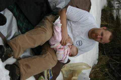 2003 Jeep TJ - A man holds a baby while sitting on a tarp in a snowy, outdoor environment.