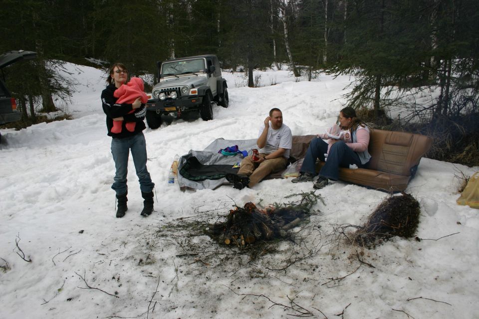 People gathered around a campfire in a snowy wooded area. A woman holds a baby, two people sit on a couch, and another person sits near a tent