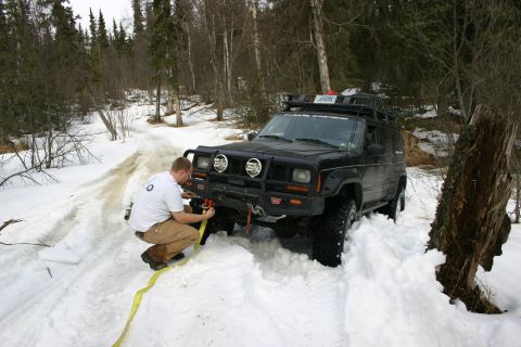 2003 Jeep TJ - Man attaches winch line to a black Jeep Cherokee XJ stuck in a snowy forest trail.