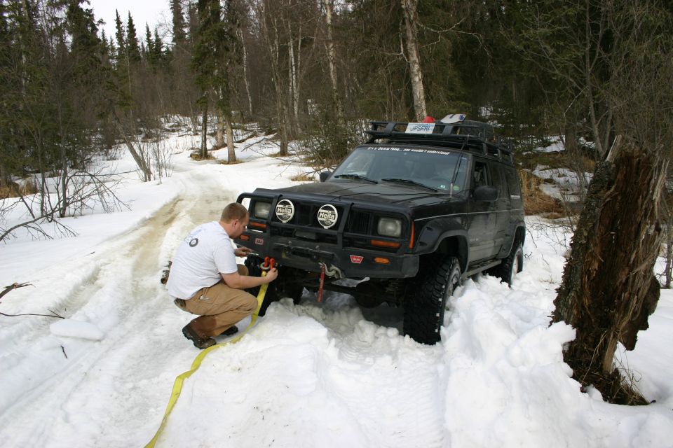 Man attaching winch line to front of black Jeep Cherokee XJ stuck in snow. Winch and roof rack visible