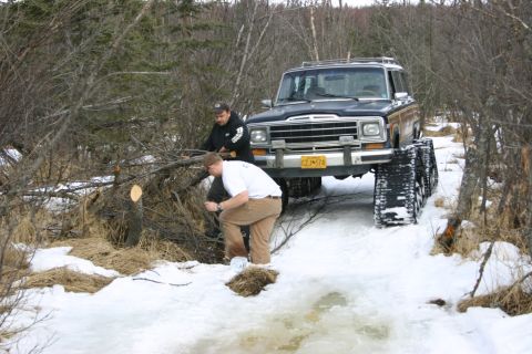2003 Jeep TJ - Jeep Cherokee equipped with tracks navigates a snowy trail while two individuals clear fallen branches.