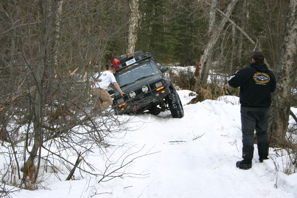 Black Jeep Cherokee XJ with roof rack stuck in snow, person pushing it