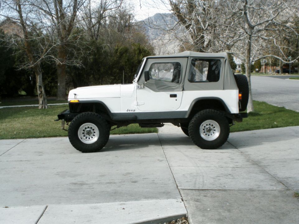 White Jeep Wrangler parked on a driveway, side view. It has a gray soft top and aftermarket wheels