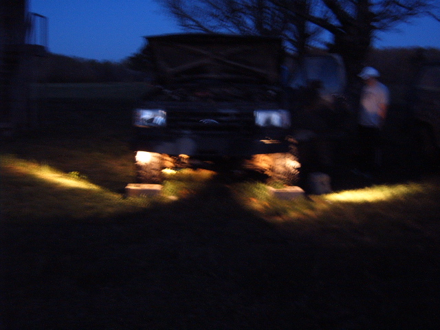 Blurry dusk photo of a Ford truck with hood up, headlights lit across grass, person standing nearby