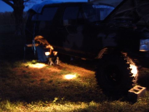1988 Ford Bronco - A truck is raised on jack stands at night, with lights casting a glow on the grass.