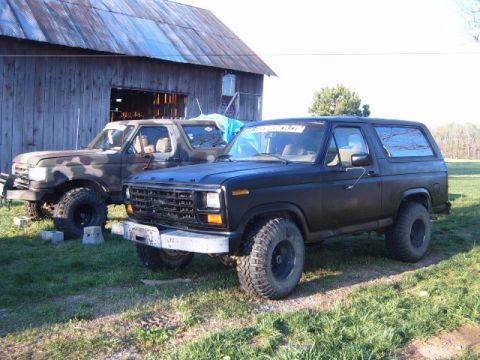 1988 Ford Bronco - Two Ford Broncos, one black and one camouflage, are parked outside a wooden barn.