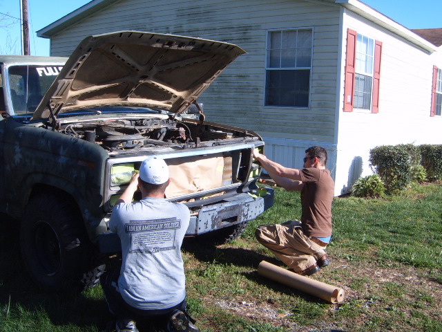 Two guys working on the front of an old Ford Bronco, hood up, near a house
