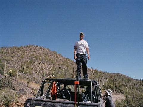 1989 Ford Bronco - Man stands atop a modified off-road vehicle in the desert, with a mountainous, cactus-filled background.