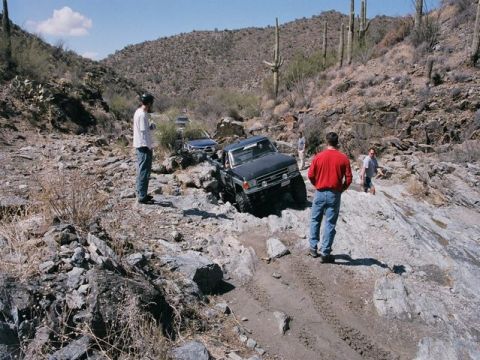 1989 Ford Bronco - Several trucks are off-roading on a rocky, desert trail with saguaro cacti in the background.