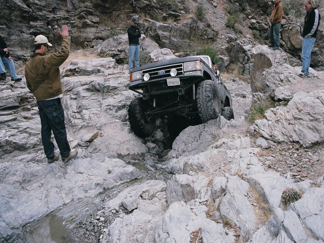 Lifted Ford Bronco crawling through a rocky canyon wash while a spotter directs it