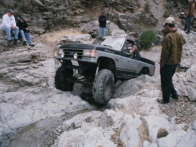 Lifted black and gray Ford 4x4 with top off crawling over boulders as spectators watch
