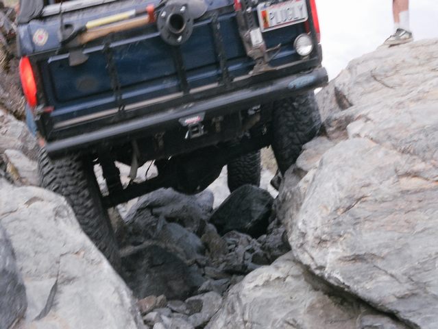 Dark blue Ford Bronco crawling up large boulders, rear view showing flexed suspension and PLUGLY plate