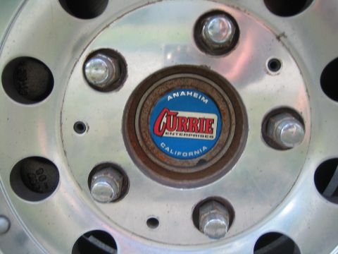 1990 Ford Bronco - Close-up of an aluminum wheel and lug nuts with a rusty hub and a blue-and-red Currie Enterprises Anaheim California center cap.