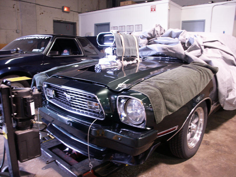 Dark green muscle car with a prominent supercharger, covered partially with a car cover and towels, on a dynamometer