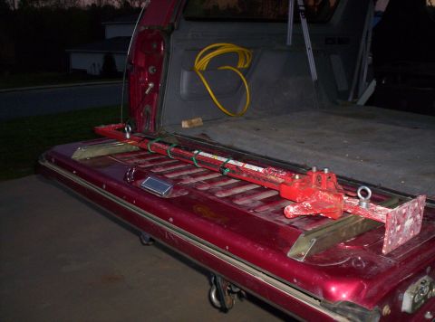 1995 Ford Bronco - A red Hi-Lift-style farm jack laid across the open maroon tailgate of a truck at dusk, secured to brass-colored mounting brackets with gree…