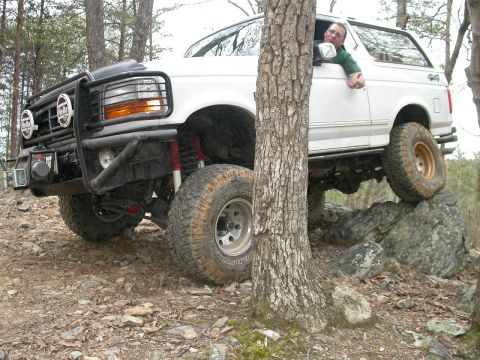 1990 Ford Bronco - A white 1990 Ford Bronco is shown off-roading, with its tires climbing over large rocks.