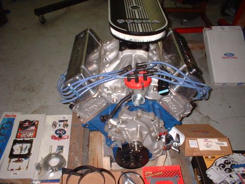 1971 Ford Mustang - A Ford Cobra engine is shown with chrome valve covers and blue spark plug wires, alongside various engine parts and boxes.