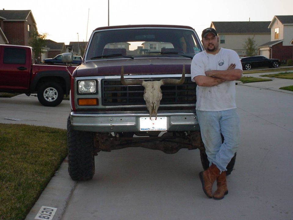 Front of maroon Ford Bronco with longhorn skull on grille; man leaning on fender in driveway