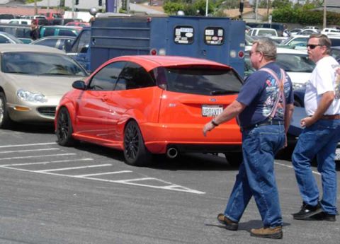1998 Ford Crown Victoria - orange Ford Focus SVT hatchback with black wheels parked beside a trailer as two men in jeans walk past.