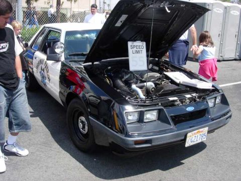 1998 Ford Crown Victoria - Fox-body Ford Mustang in black-and-white Highway Patrol livery sits hood-up with a Speed Limit 55 sign on the engine.