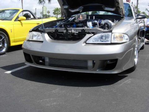 1998 Ford Crown Victoria - Front of a silver SN95 Mustang with custom bumper, front-mount intercooler, and turbo plumbing under the raised hood.