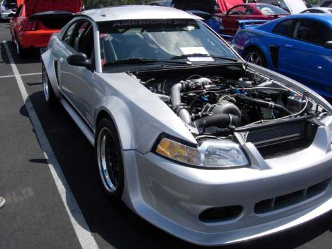 1998 Ford Crown Victoria - silver New Edge Ford Mustang with a custom turbo engine setup sits hood-up among cars at a show.
