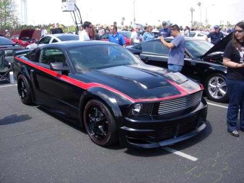 1998 Ford Crown Victoria - black S197 Ford Mustang with a red pinstripe, custom body kit, and black wheels parked at a busy car show.