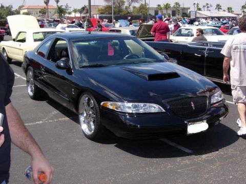 1998 Ford Crown Victoria - glossy black Lincoln coupe with chrome wheels and a hood scoop sits among other classics in a sunlit car-show parking lot.