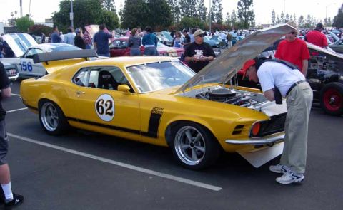1998 Ford Crown Victoria - yellow Boss 302 Mustang race car wearing number 62 has its hood open as a man inspects the engine at a packed car show.